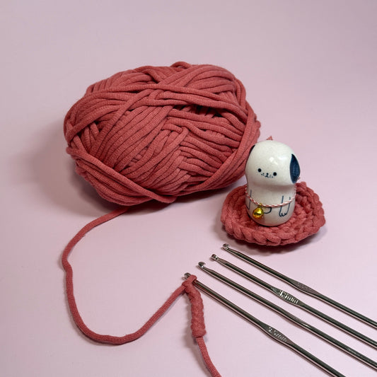 Ball of pink yarn with knitting needles and a small white figurine on a light pink background